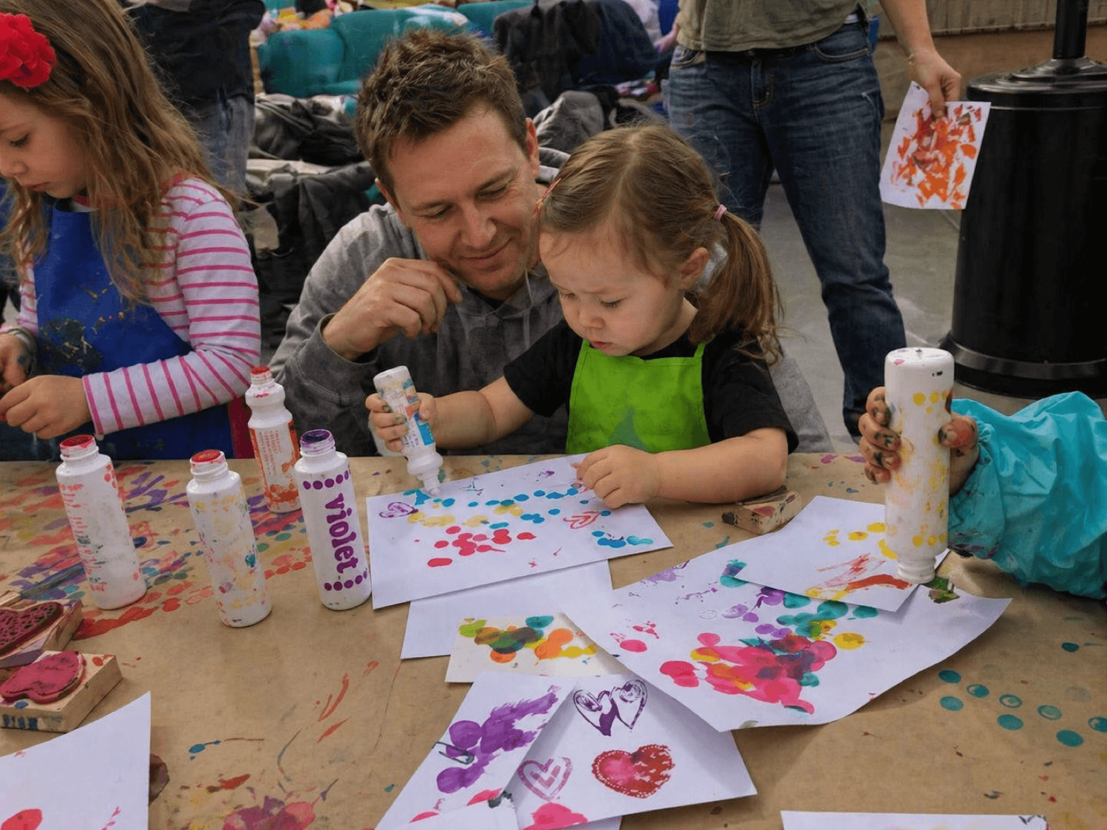 A team painting together at a long outdoor table at golden hour — finished canvases on easels